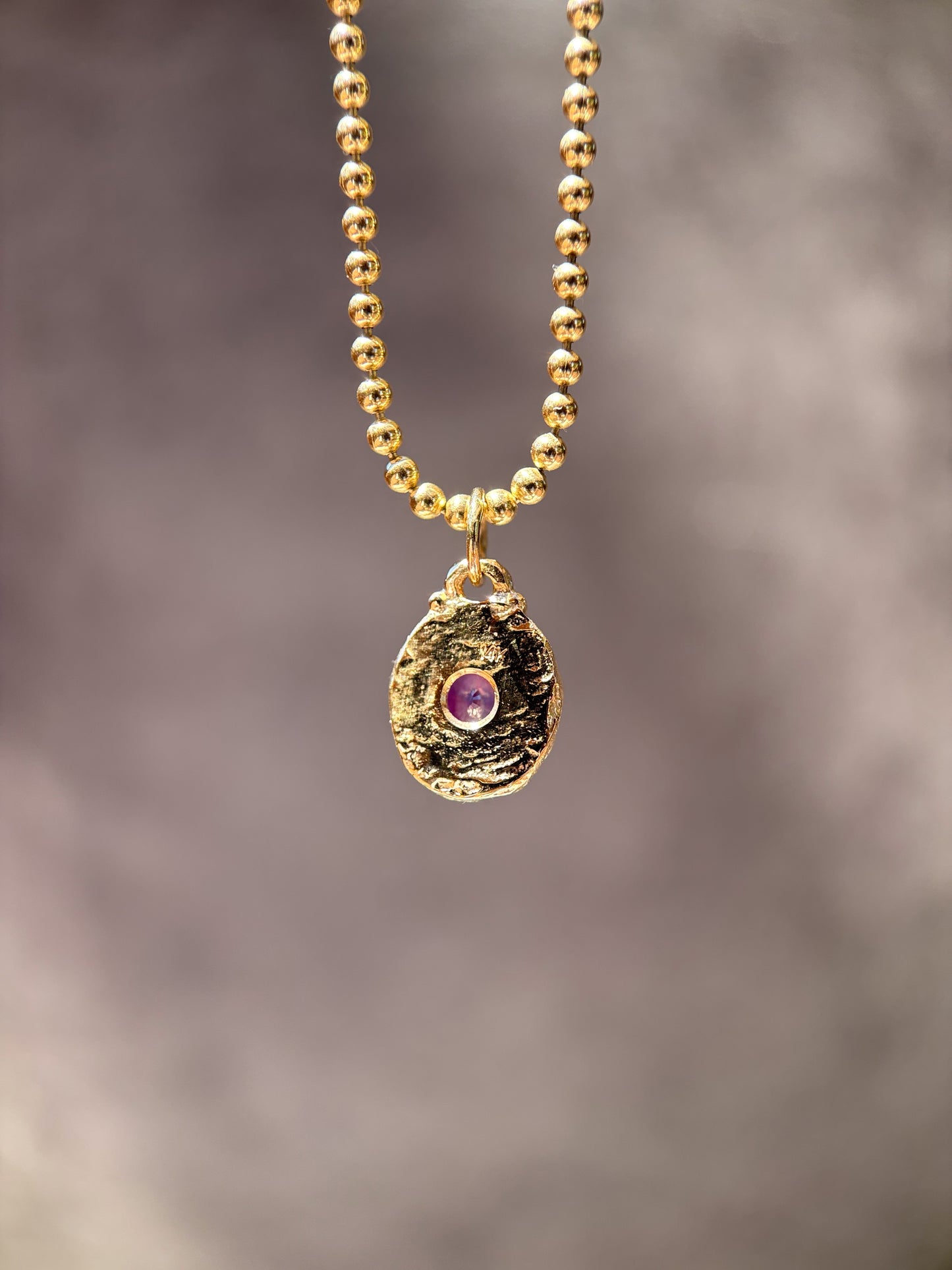 A gold beaded necklace with a textured, organic-shaped gold pendant featuring a small purple gemstone in the center, set against a soft, blurred gray background.