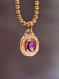 A gold ball-chain necklace with a textured gold pendant featuring a central oval purple gemstone, shown against a blurred dark brown background.