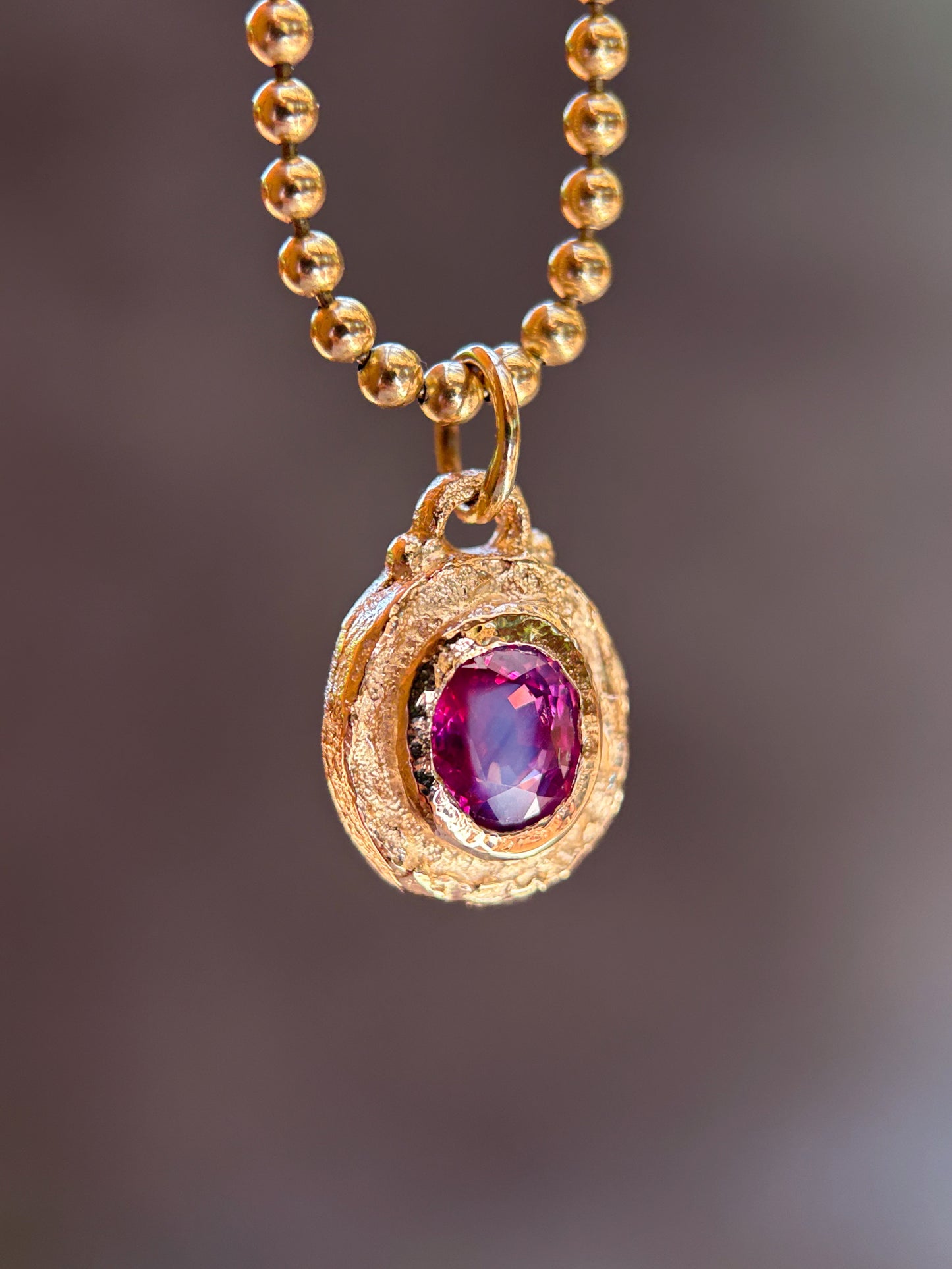 A gold ball-chain necklace with a textured gold pendant featuring a central oval purple gemstone, shown against a blurred dark brown background.