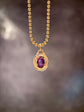 A gold beaded chain necklace with an oval pendant featuring a deep purple gemstone surrounded by a textured gold border, set against a dark, blurred background.