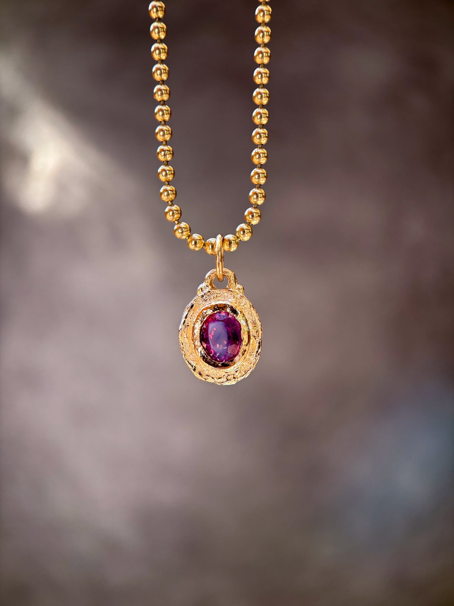 A gold beaded chain necklace with an oval pendant featuring a deep purple gemstone surrounded by a textured gold border, set against a dark, blurred background.