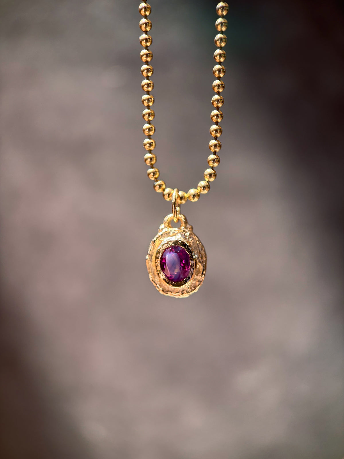 A gold ball chain necklace with an oval pendant featuring a textured gold frame and a deep purple oval gemstone, shown against a soft, blurred gray background.