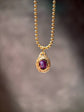 A gold ball chain necklace with an oval pendant featuring a textured gold frame and a deep purple oval gemstone, shown against a soft, blurred gray background.