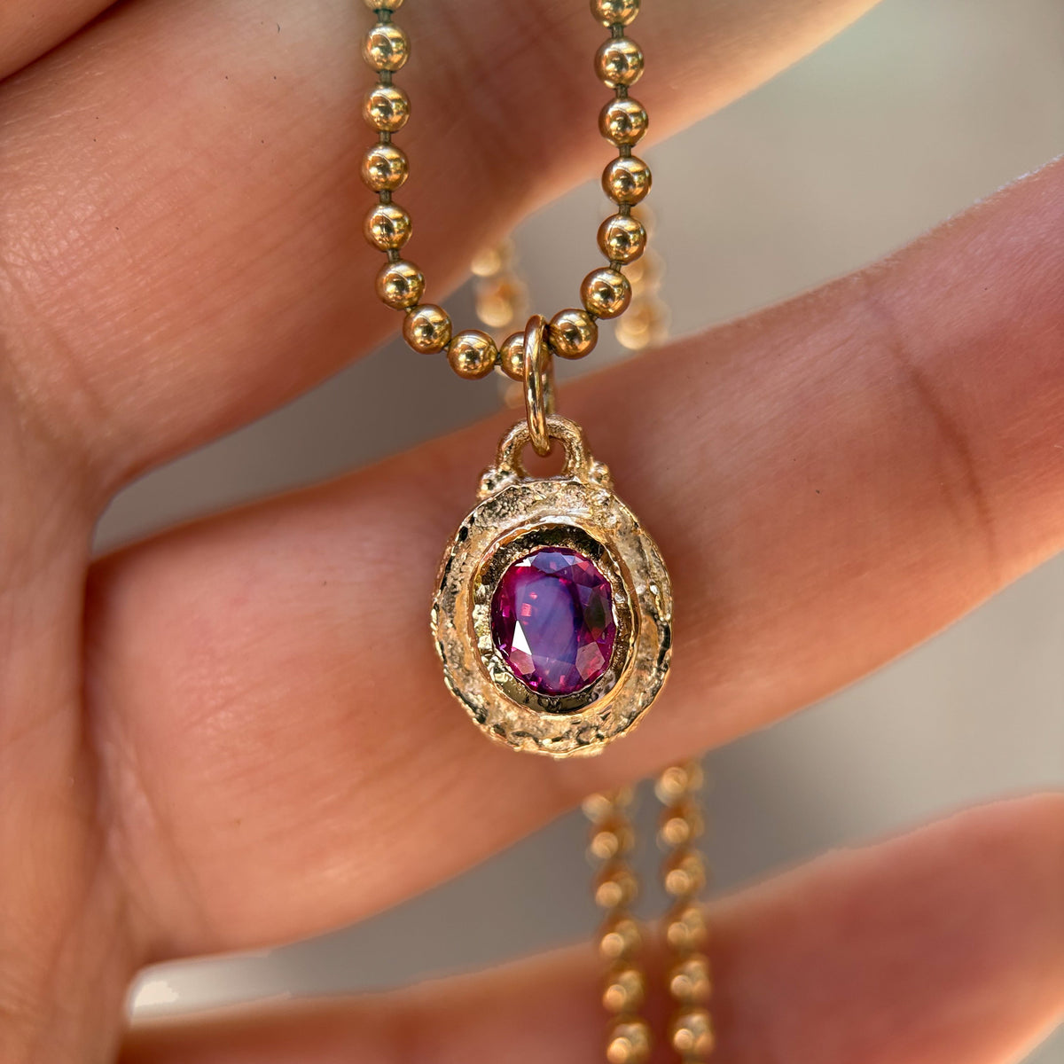 A gold ball chain necklace with a textured gold pendant featuring a purple oval gemstone, held in a person's hand.