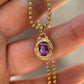 A gold ball chain necklace with a textured gold pendant featuring a purple oval gemstone, held in a person's hand.