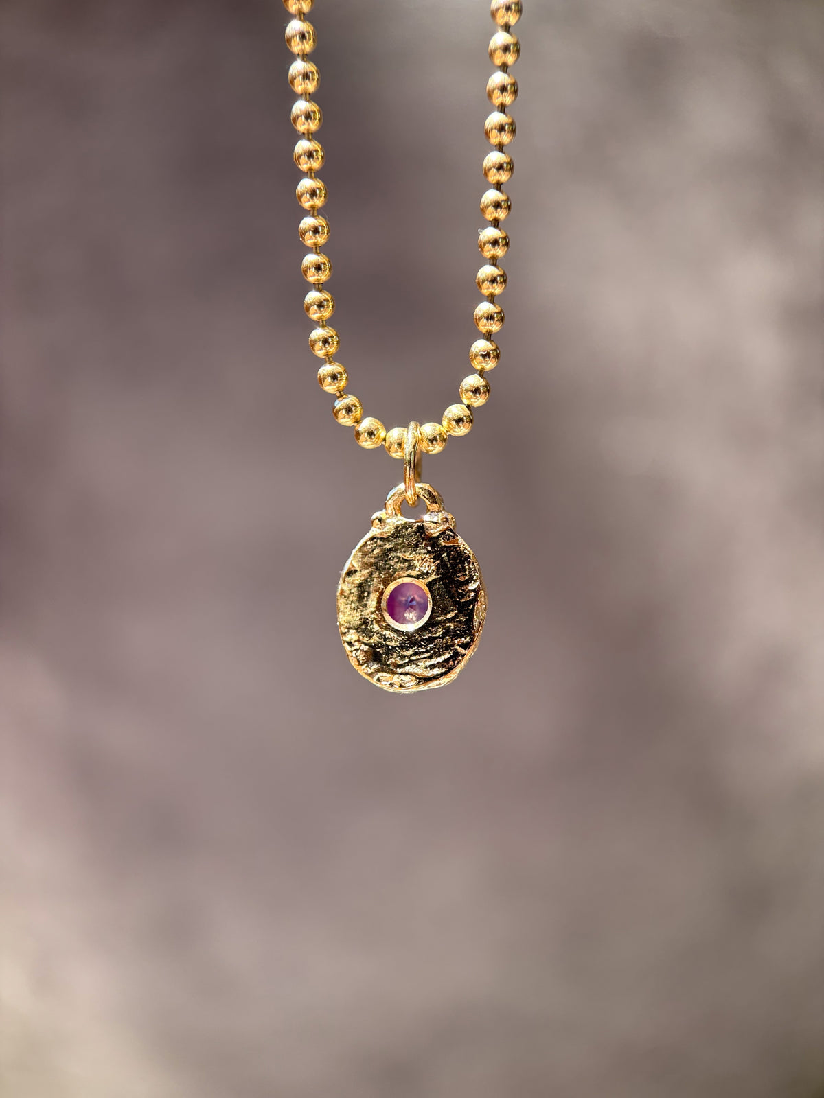 A gold beaded necklace with a textured, organic-shaped gold pendant featuring a small purple gemstone in the center, set against a soft, blurred gray background.