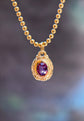 A gold ball-chain necklace with a textured gold pendant featuring an oval purple gemstone, shown against a blurred dark background.