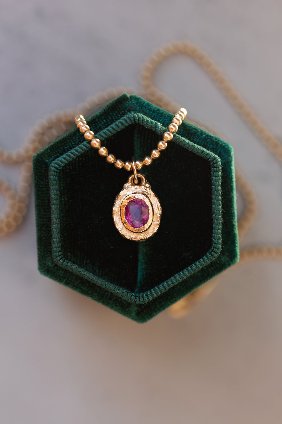 A gold beaded necklace with an oval pendant featuring a purple gemstone surrounded by small clear stones, displayed on a dark green velvet hexagonal jewelry box.