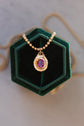 A gold beaded necklace with an oval pendant featuring a purple gemstone surrounded by small clear stones, displayed on a dark green velvet hexagonal jewelry box.