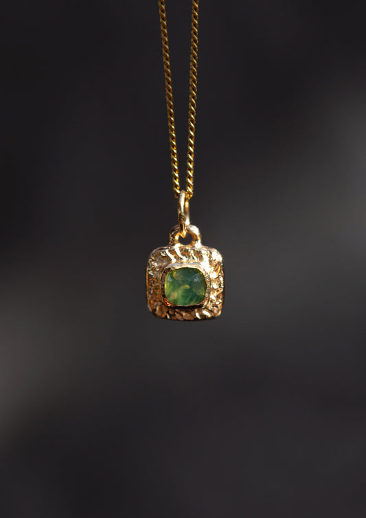 A gold pendant necklace featuring a textured gold bezel setting around a square-cut green gemstone, suspended from a gold chain against a dark, blurred background.
