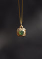 A gold pendant necklace featuring a textured gold bezel setting around a square-cut green gemstone, suspended from a gold chain against a dark, blurred background.