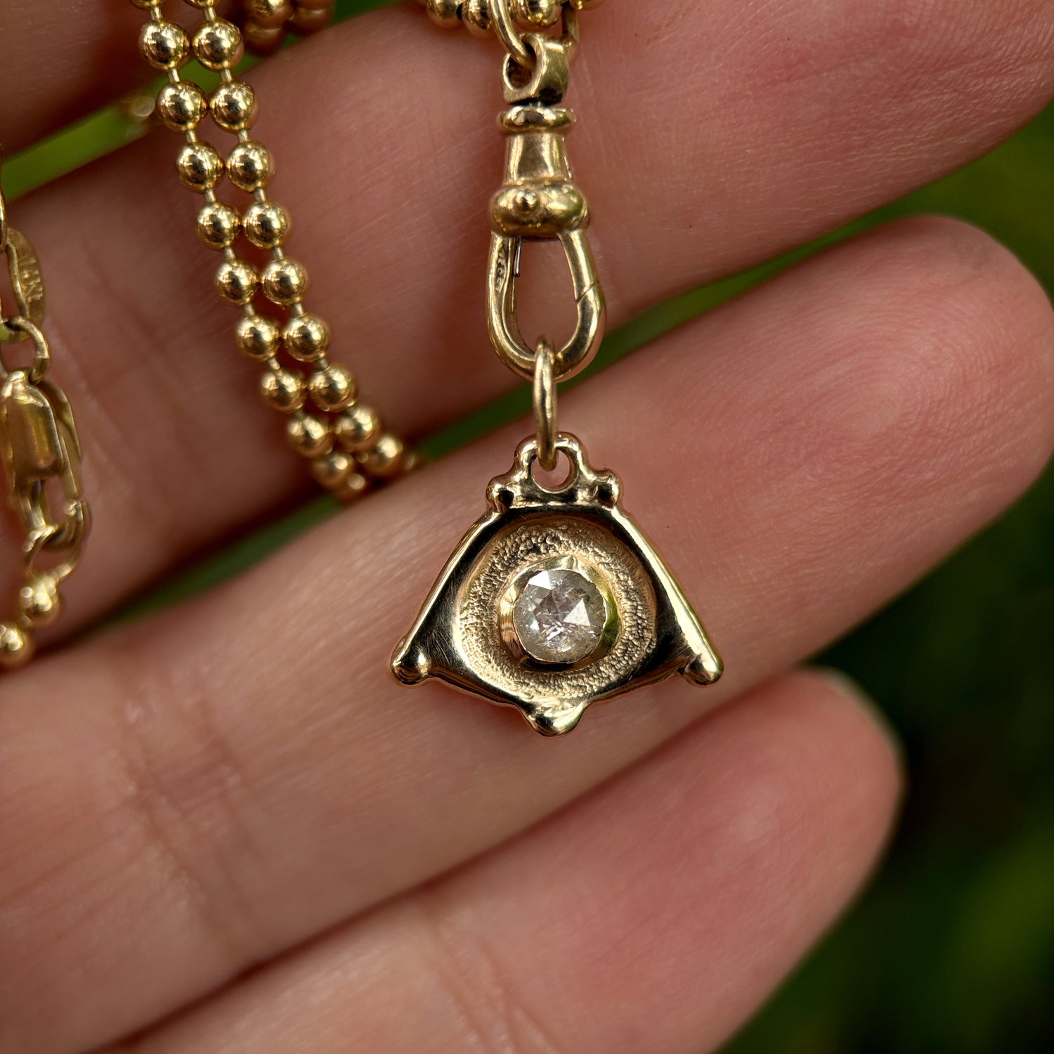 Gold necklace with a pendant featuring a diamond held between fingers against a blurred green background