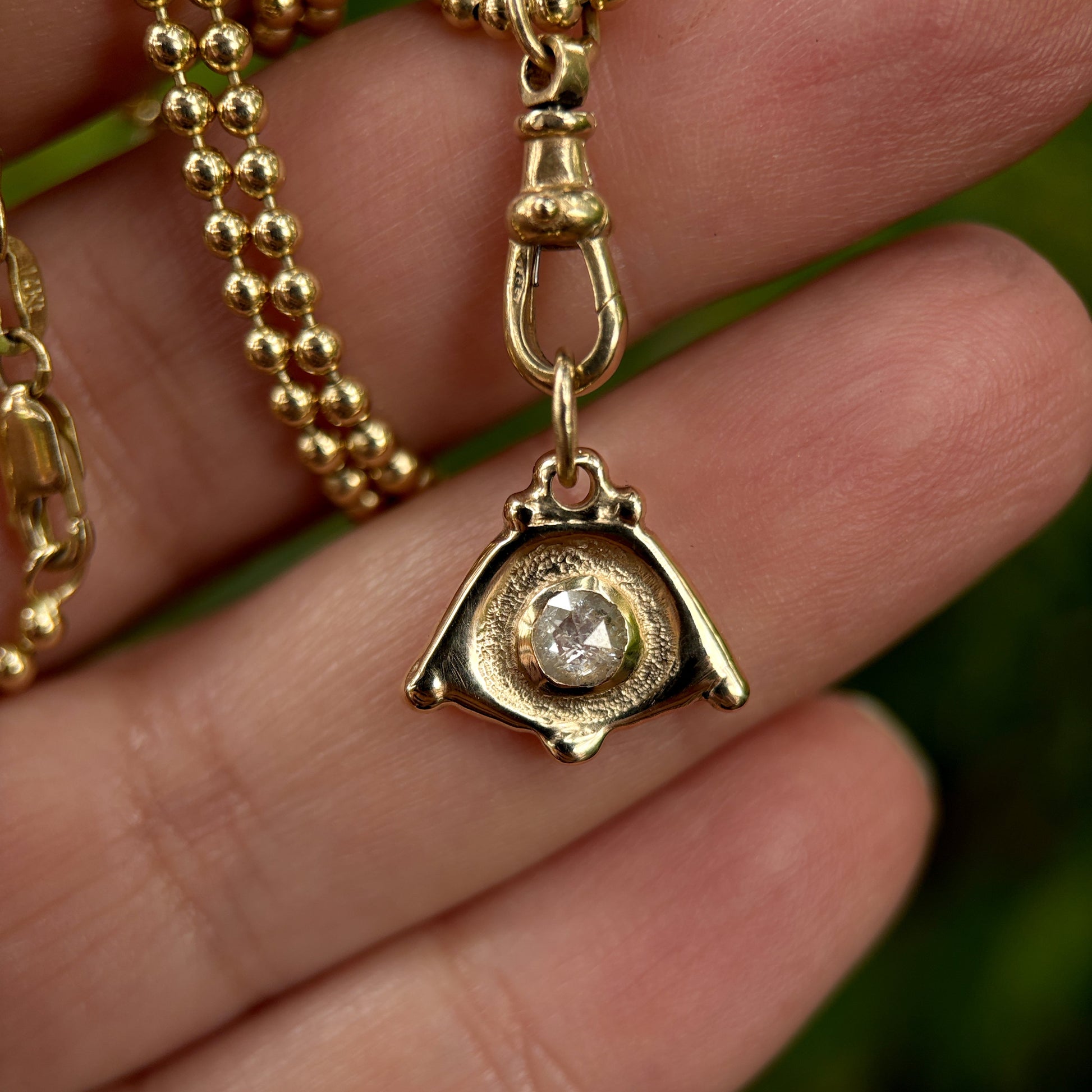 Gold necklace with a pendant featuring a diamond held between fingers against a blurred green background