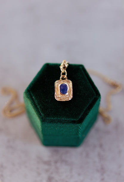 Gold pendant with a blue gemstone on a green velvet stand against a neutral background