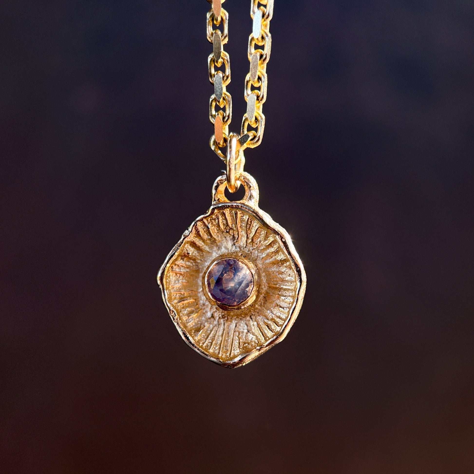 Gold necklace with a round pendant featuring a purple gemstone on a dark background