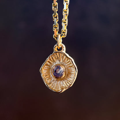 Gold necklace with a round pendant featuring a purple gemstone on a dark background