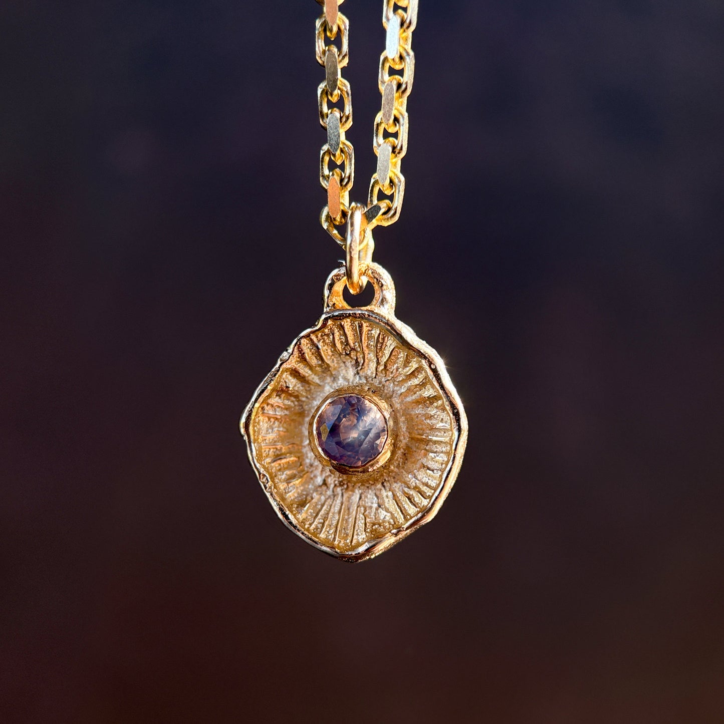 Gold necklace with a round pendant featuring a purple gemstone on a dark background