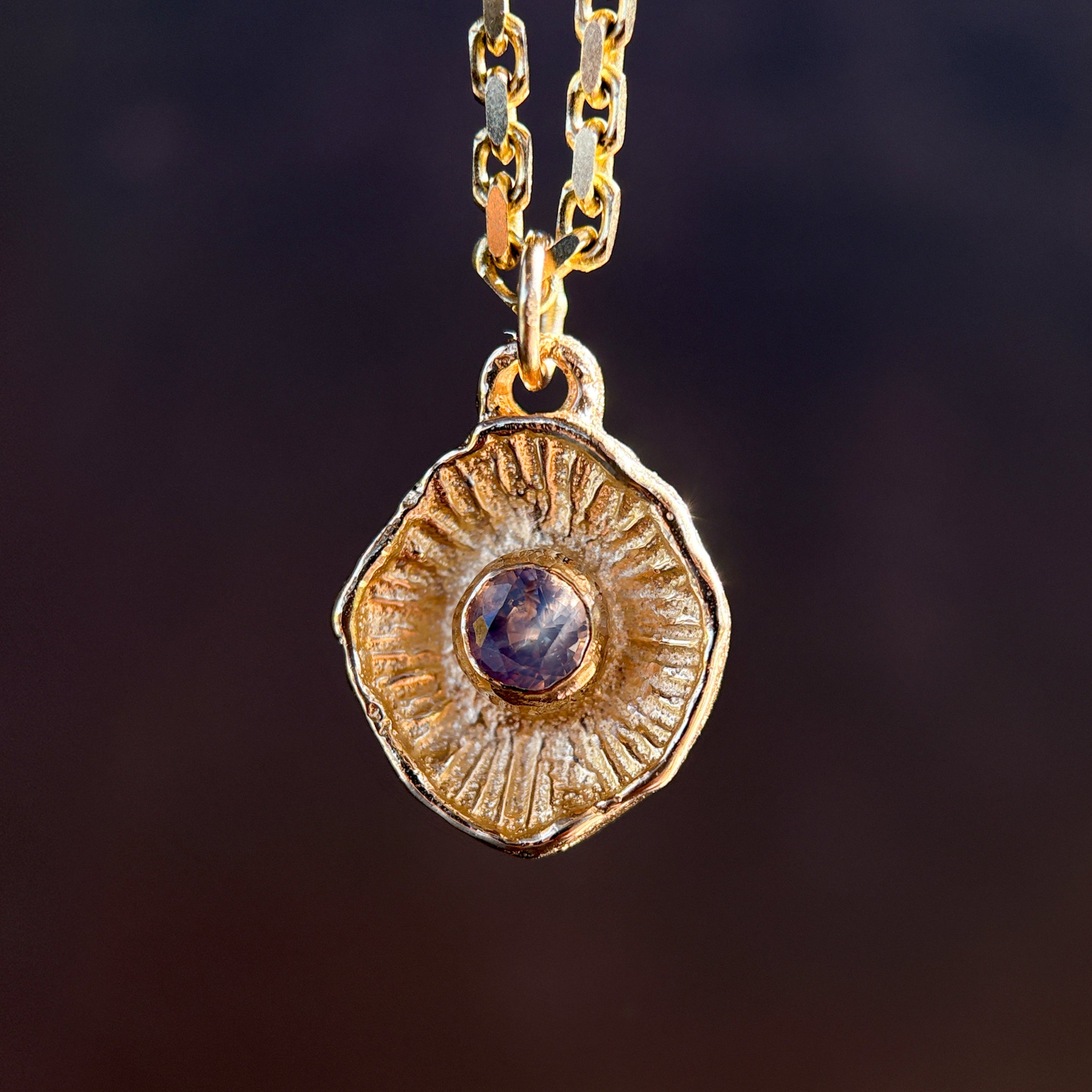 Gold necklace with a round pendant featuring a purple gemstone on a dark background