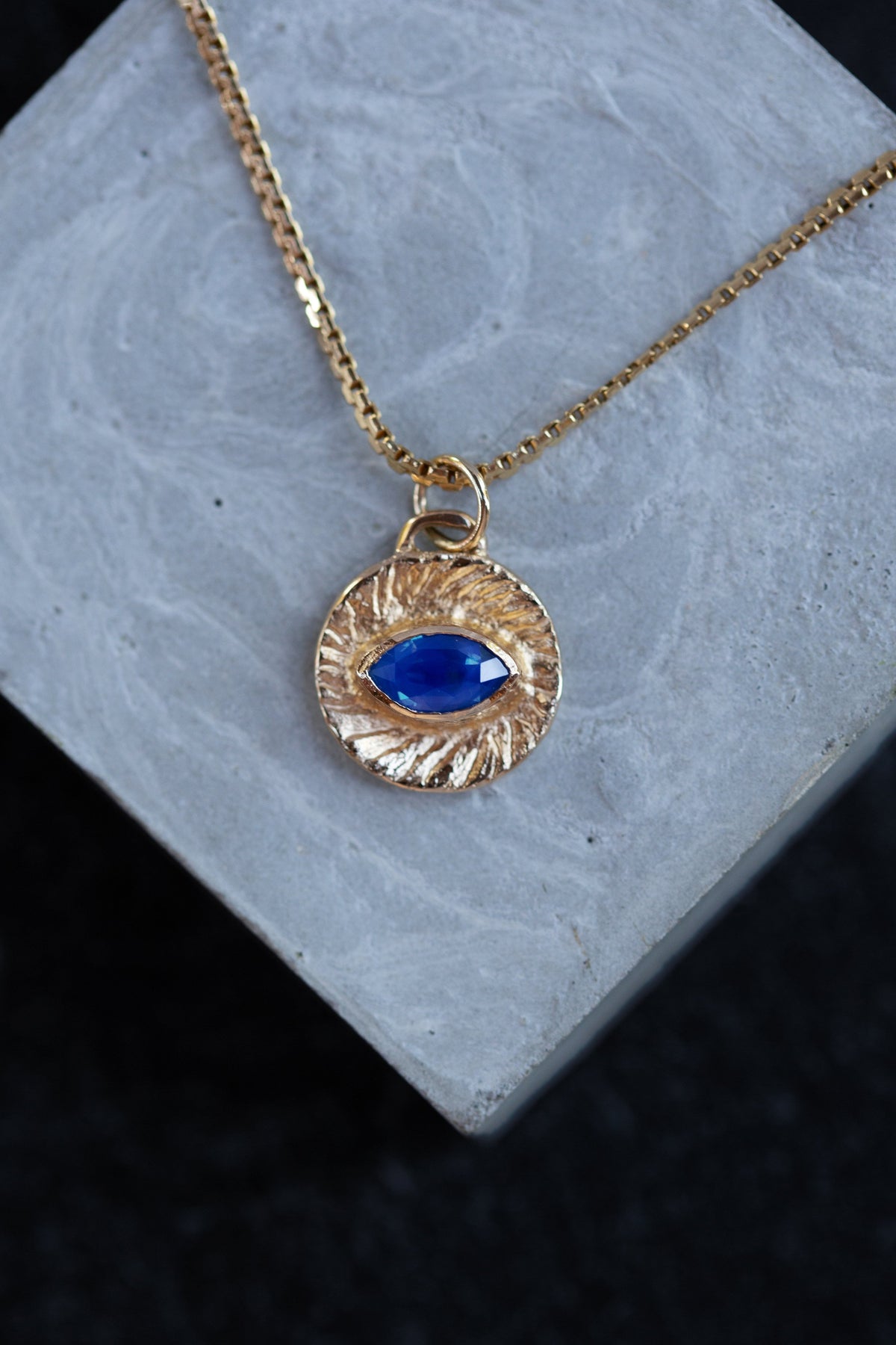 A gold chain necklace with a textured circular pendant featuring a blue eye motif, displayed on a light gray stone block against a dark background.