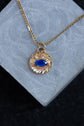 A gold chain necklace with a textured circular pendant featuring a blue eye motif, displayed on a light gray stone block against a dark background.