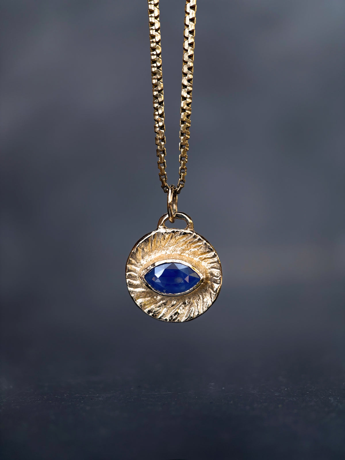 A gold-toned necklace with a textured circular pendant featuring a central oval blue gemstone, suspended against a dark gray background.