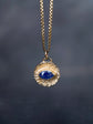 A gold-toned necklace with a textured circular pendant featuring a central oval blue gemstone, suspended against a dark gray background.