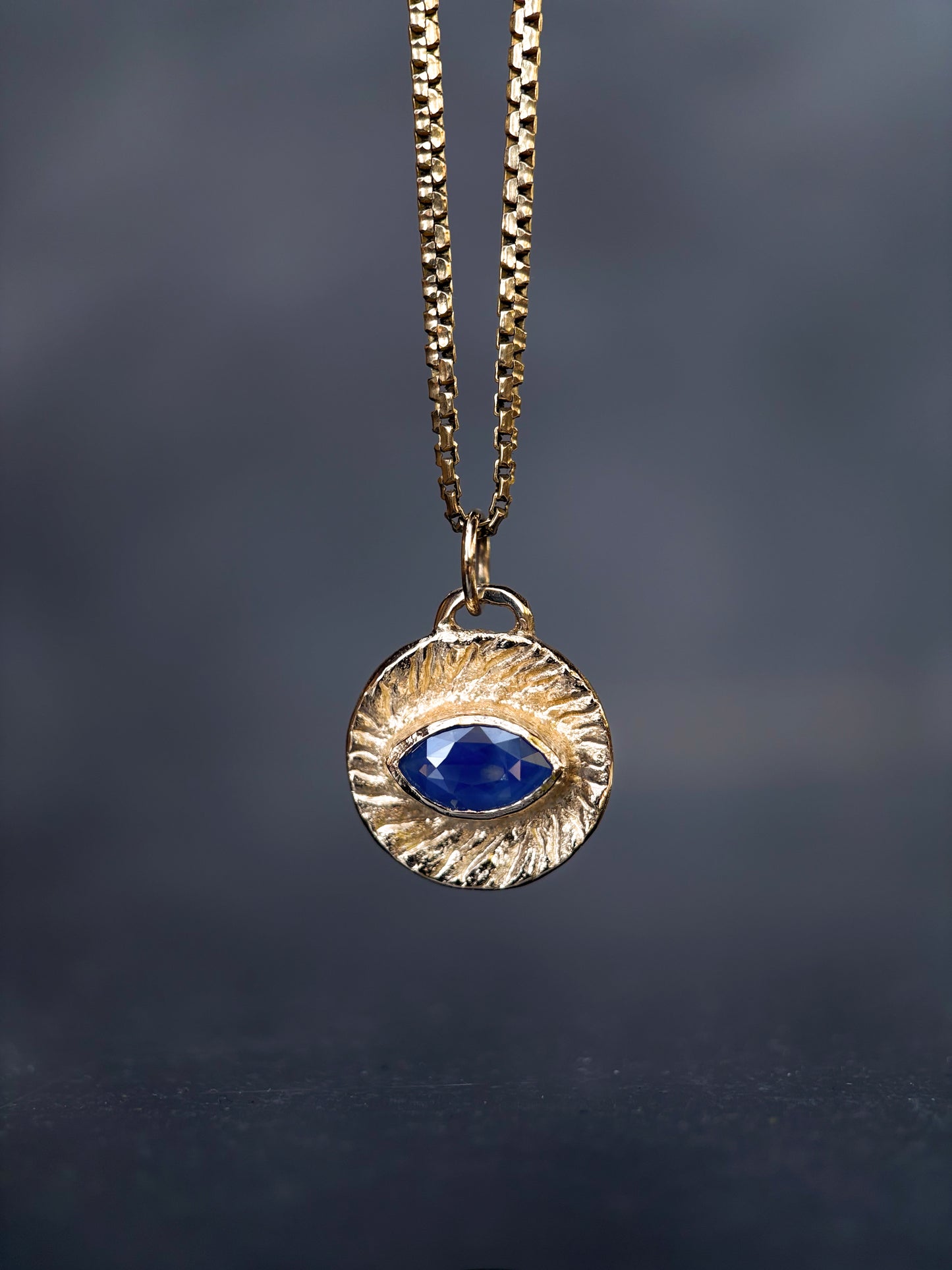 A gold-toned necklace with a textured circular pendant featuring a central oval blue gemstone, suspended against a dark gray background.