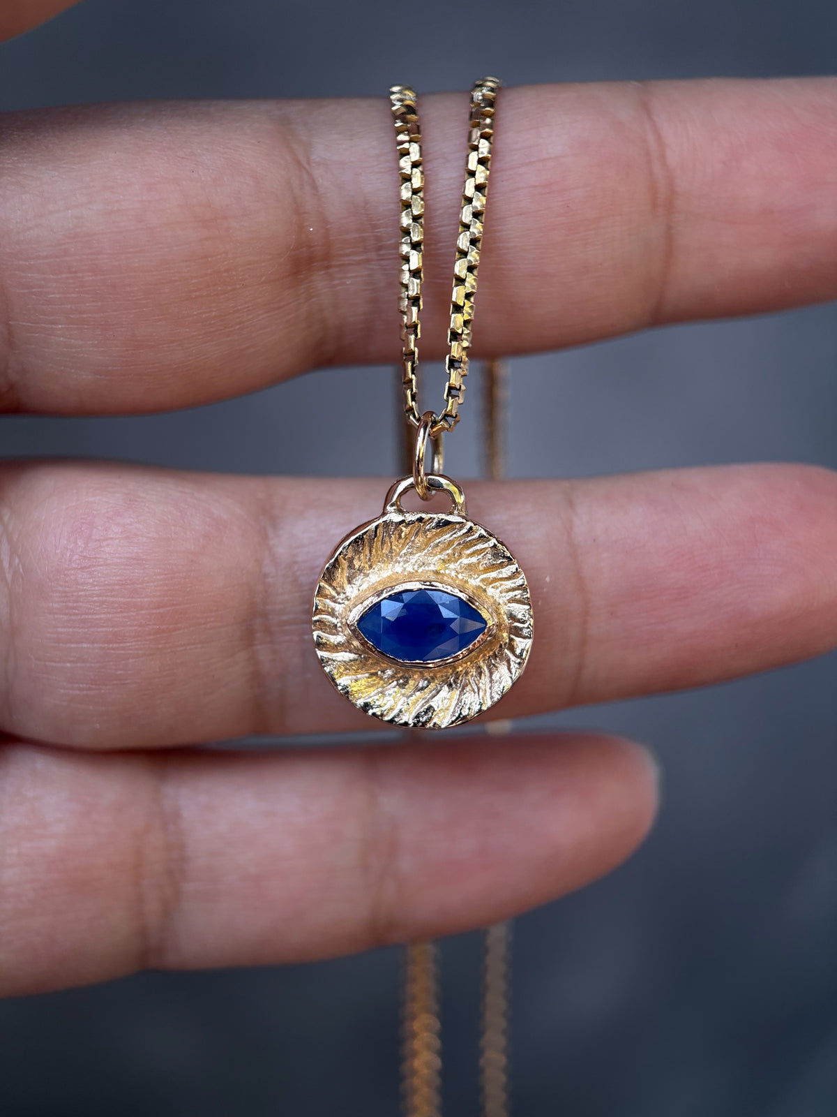 A gold necklace with a textured circular pendant featuring a central blue teardrop gemstone, held between fingers against a dark gray background.