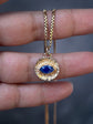 A gold necklace with a textured circular pendant featuring a central blue teardrop gemstone, held between fingers against a dark gray background.