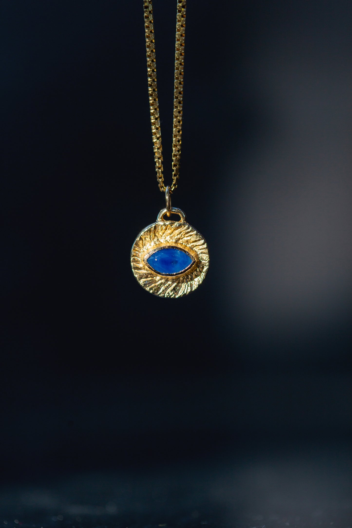 A gold pendant necklace featuring a textured circular medallion with a central blue oval stone, suspended against a dark, blurred background.