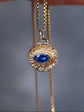 A gold-toned necklace with a textured circular pendant featuring a deep blue teardrop gemstone, held by a hand against a blurred gray background.