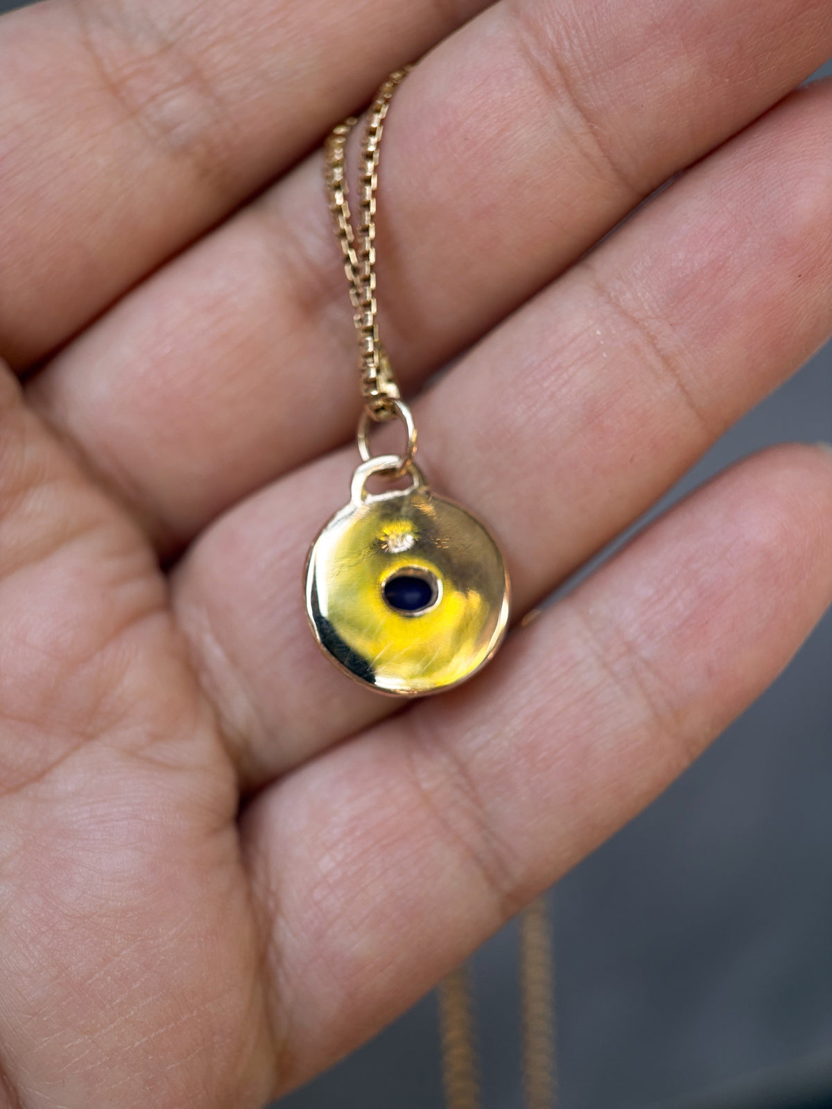A gold-toned necklace with a circular pendant featuring a central black dot, held in a person's hand.