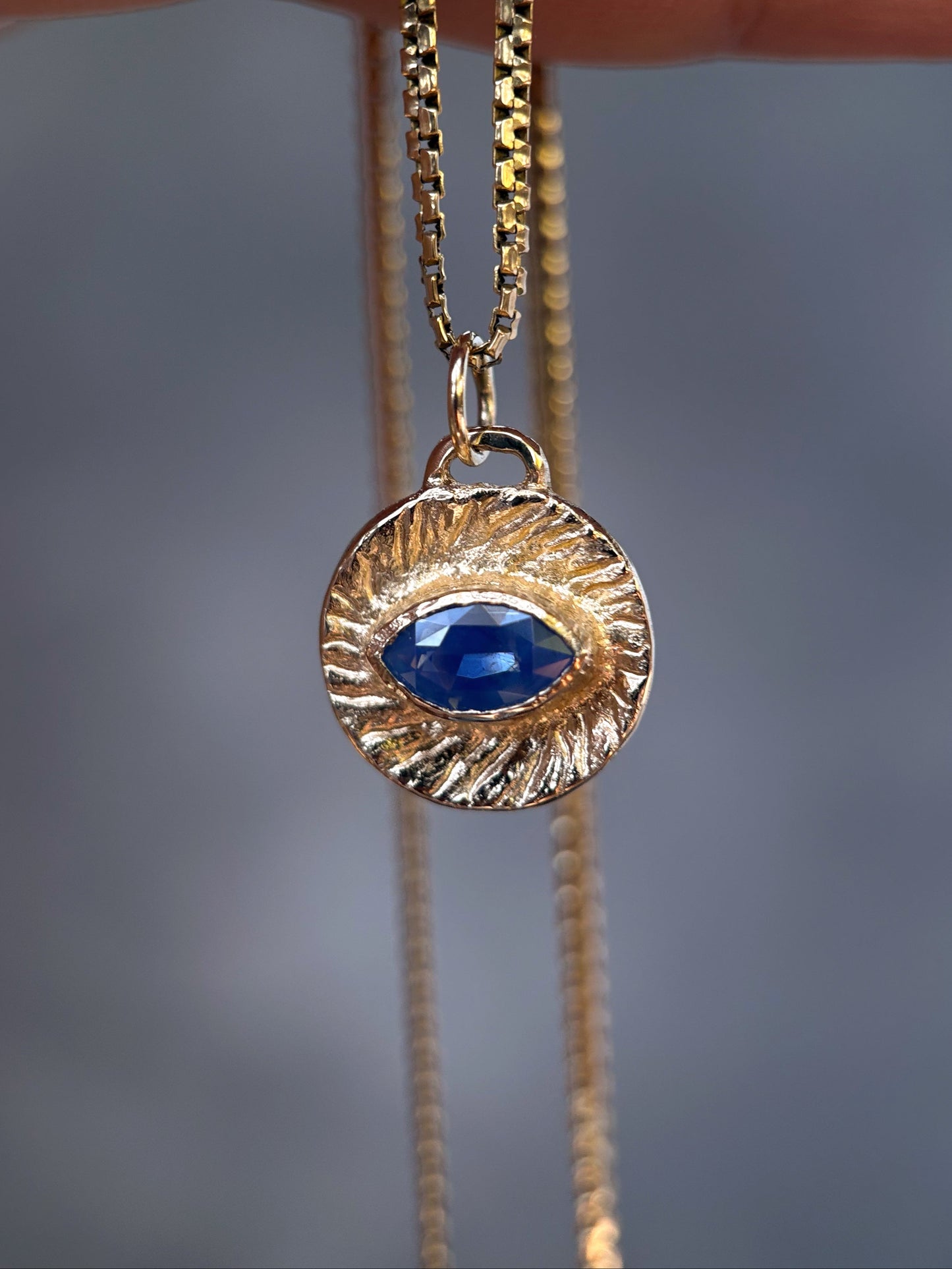 A gold-toned necklace with a textured circular pendant featuring a deep blue teardrop gemstone, held by a hand against a blurred gray background.