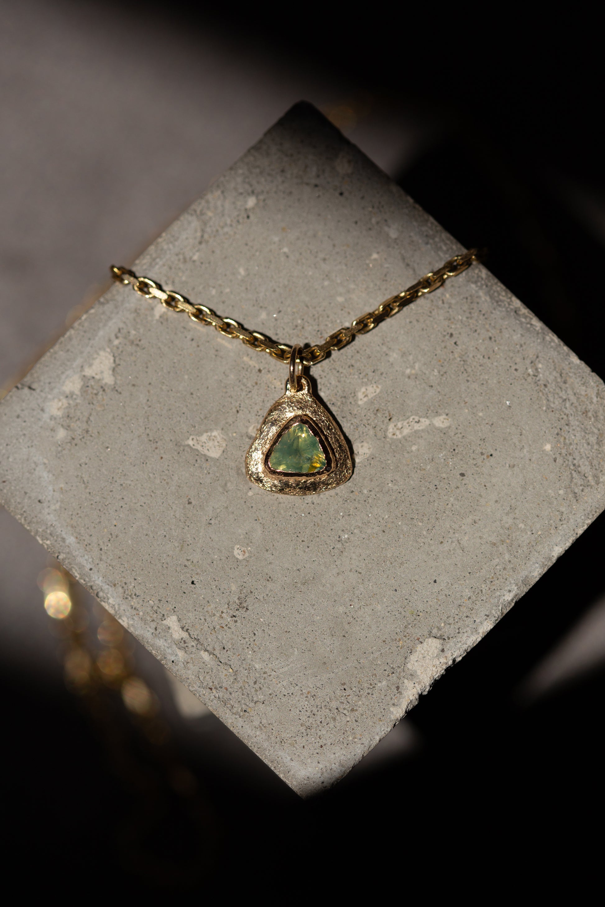 A gold chain necklace with a triangular pendant featuring a green iridescent stone, displayed on a textured gray stone block against a dark background.