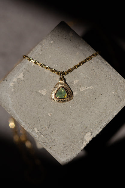 A gold chain necklace with a triangular pendant featuring a green iridescent stone, displayed on a textured gray stone block against a dark background.