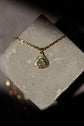 A gold chain necklace with a triangular pendant featuring a green iridescent stone, displayed on a textured gray stone block against a dark background.