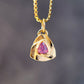 A gold pendant necklace featuring a textured triangular gold charm with a central pink teardrop gemstone, suspended from a gold box chain against a dark gray blurred background.