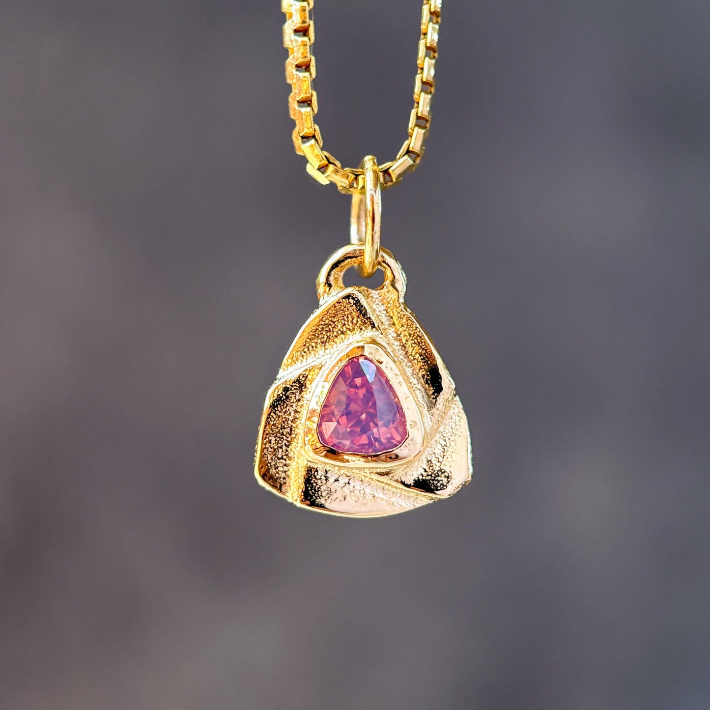 A gold pendant necklace featuring a textured triangular gold charm with a central pink teardrop gemstone, suspended from a gold box chain against a dark gray blurred background.