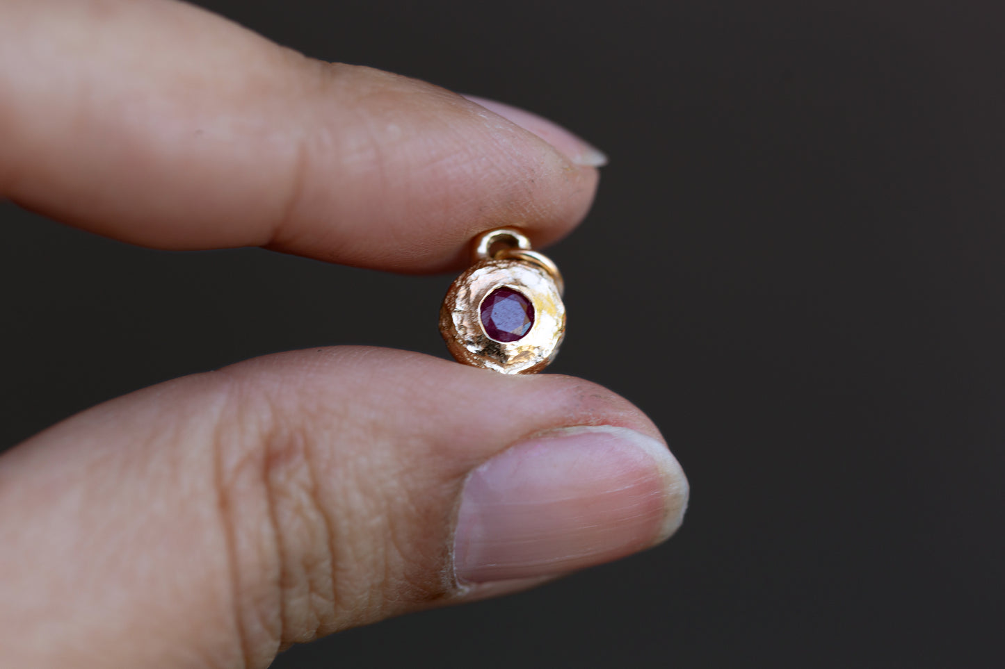 Gold pendant with a red gemstone held between fingers against a dark background