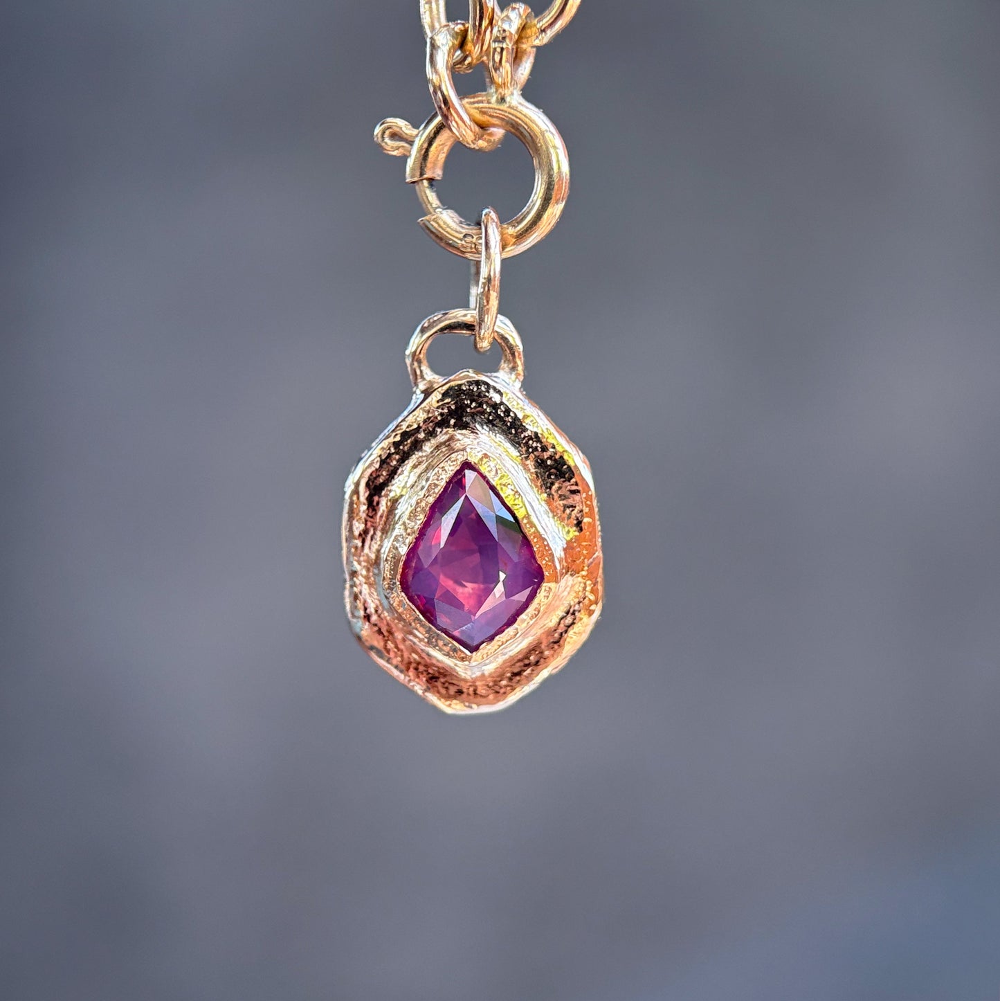 A close-up of a gold chain necklace with a textured, organic-shaped gold pendant holding a faceted purple gemstone, set against a blurred gray background.