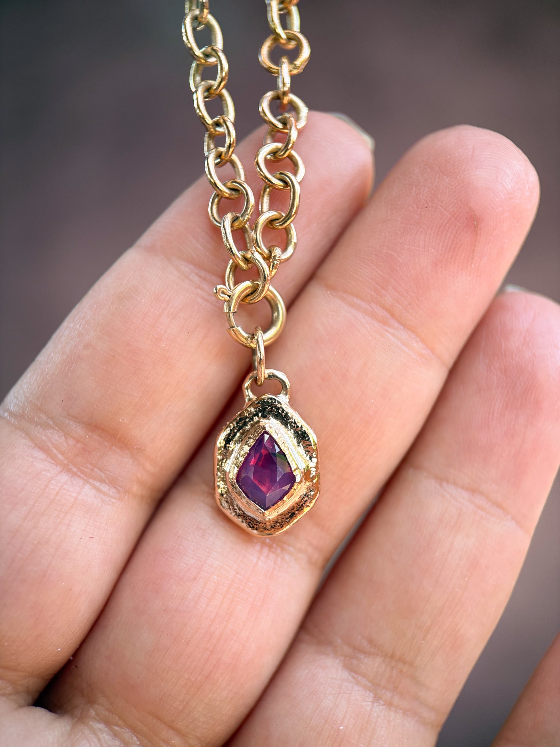 A gold chain necklace with a teardrop-shaped pendant featuring a faceted purple gemstone, held in a person's hand.