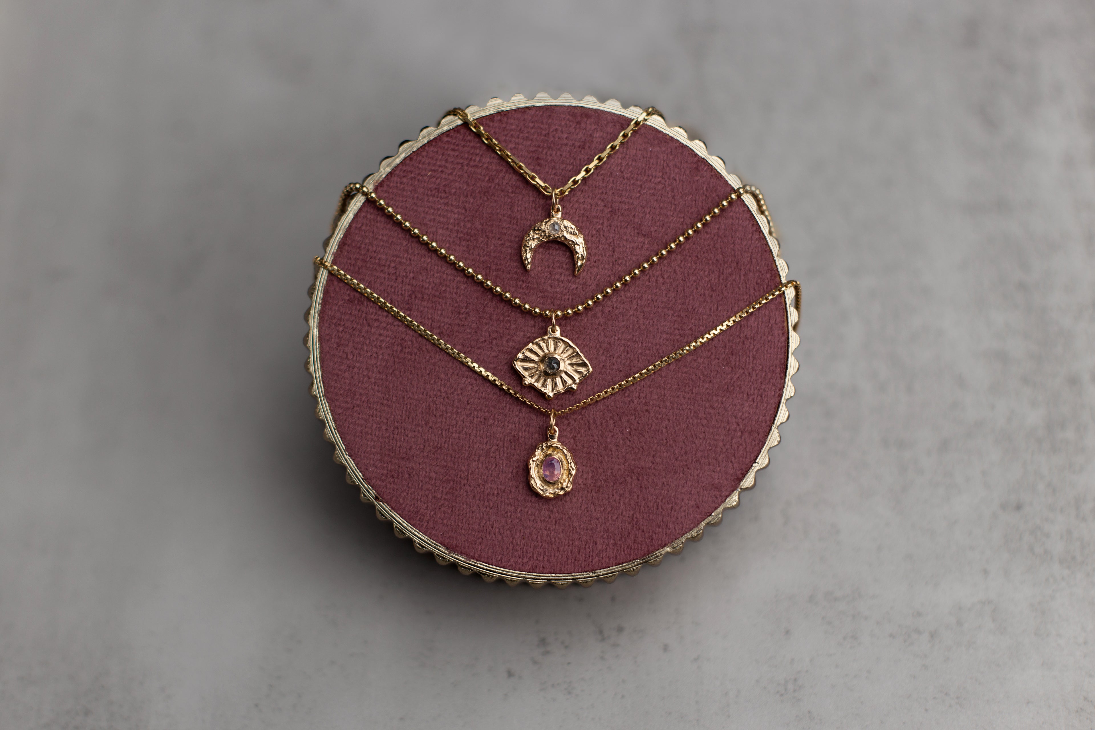 Three gold necklaces with pendants on a round purple cushion against a gray background